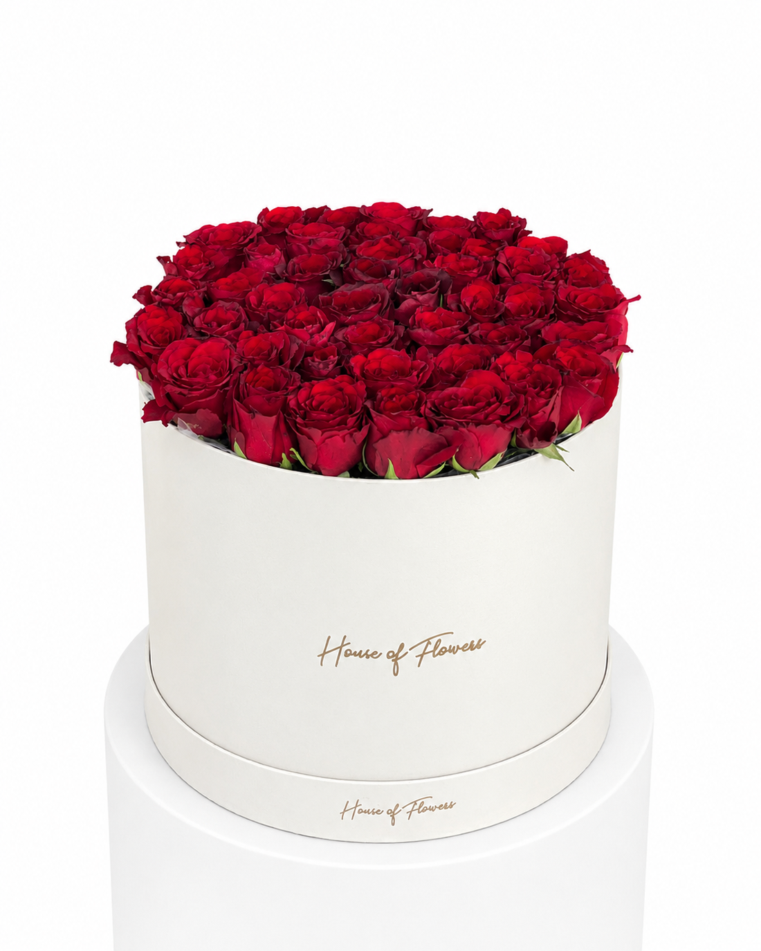 A round white flower box filled with a dense arrangement of deep red roses, displayed on a white pedestal against a white background. The box features the brand name &