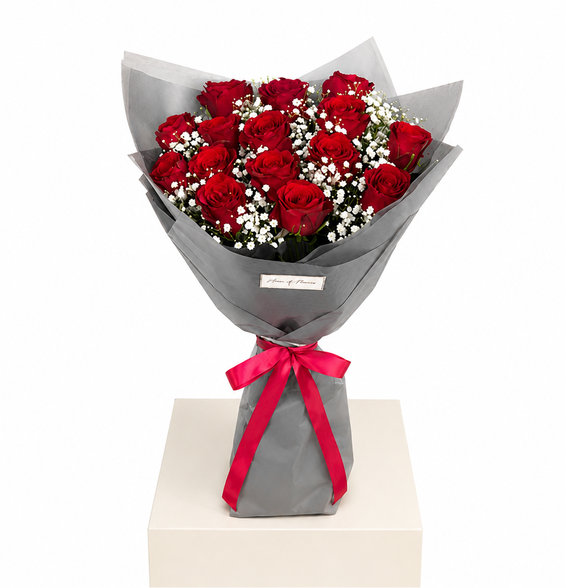A bouquet of 12 red roses wrapped in gray paper with a bright red ribbon bow, displayed on a white pedestal against a white background.
