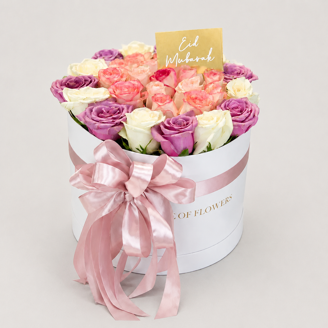A round white gift box filled with pink, peach, cream, and purple roses, tied with a large pale pink satin bow. A small gold card reading &