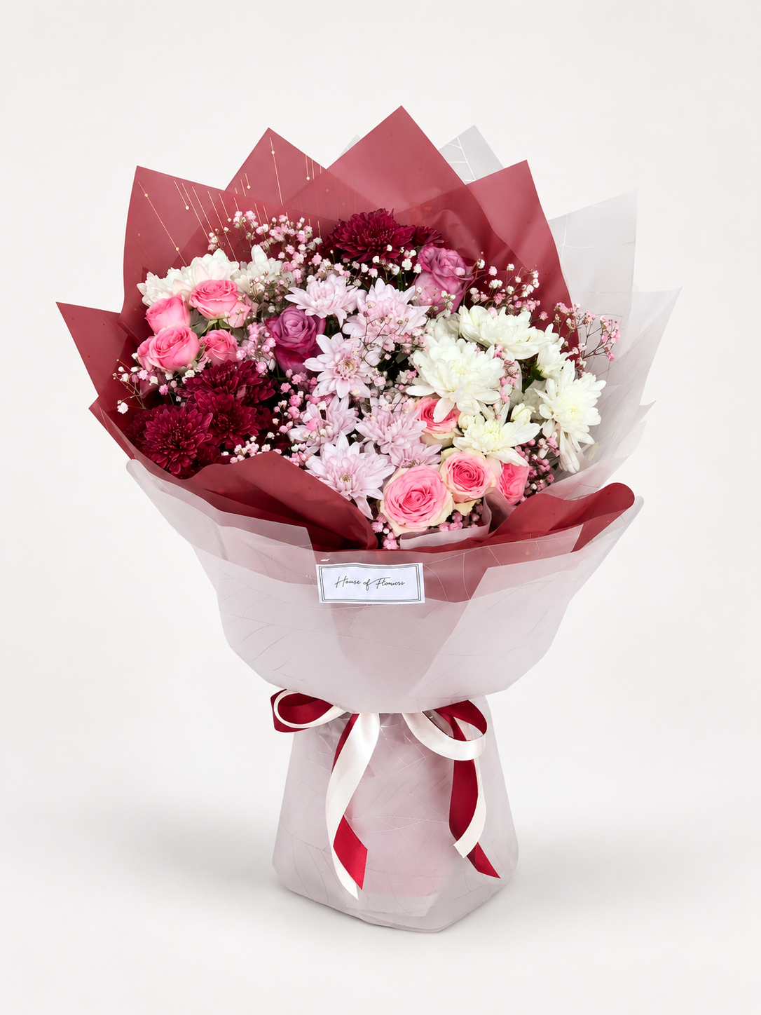 A floral bouquet wrapped in translucent pink paper with dark red tissue paper at the top, tied with a red and white striped ribbon. The bouquet contains pink and burgundy roses, white carnations, and baby&