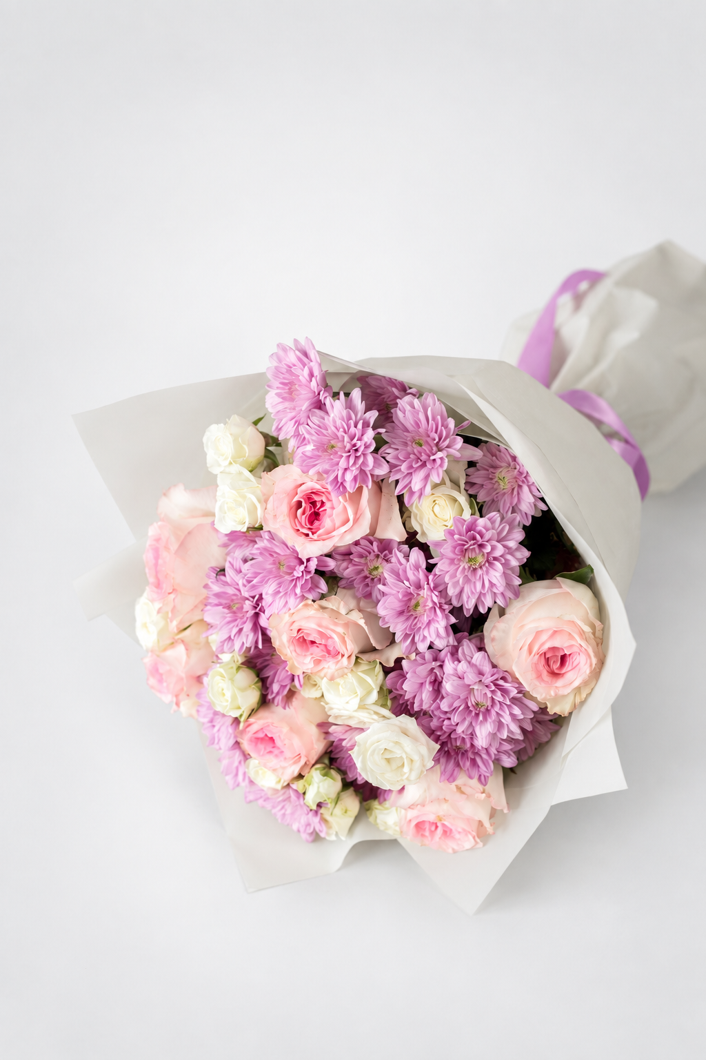 A bouquet of pink and purple flowers wrapped in white paper with a light purple ribbon, photographed on a clean white background.