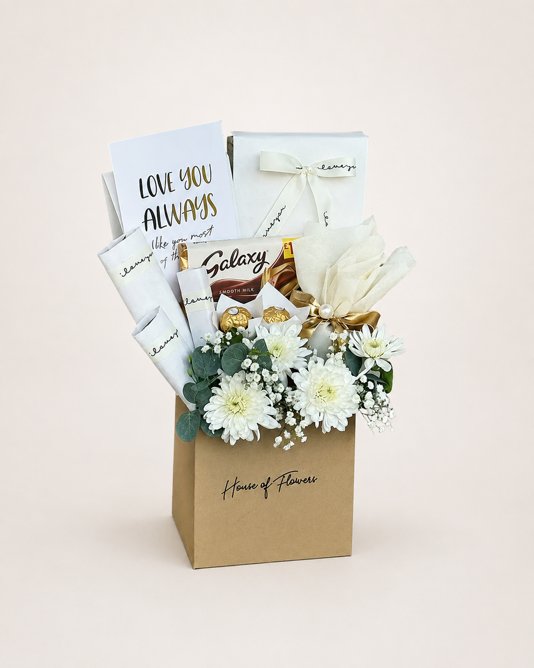 A gourmet gift box from House of Flowers, featuring a brown kraft paper bag filled with white flowers, chocolates, and gift cards, set against a solid light beige background.