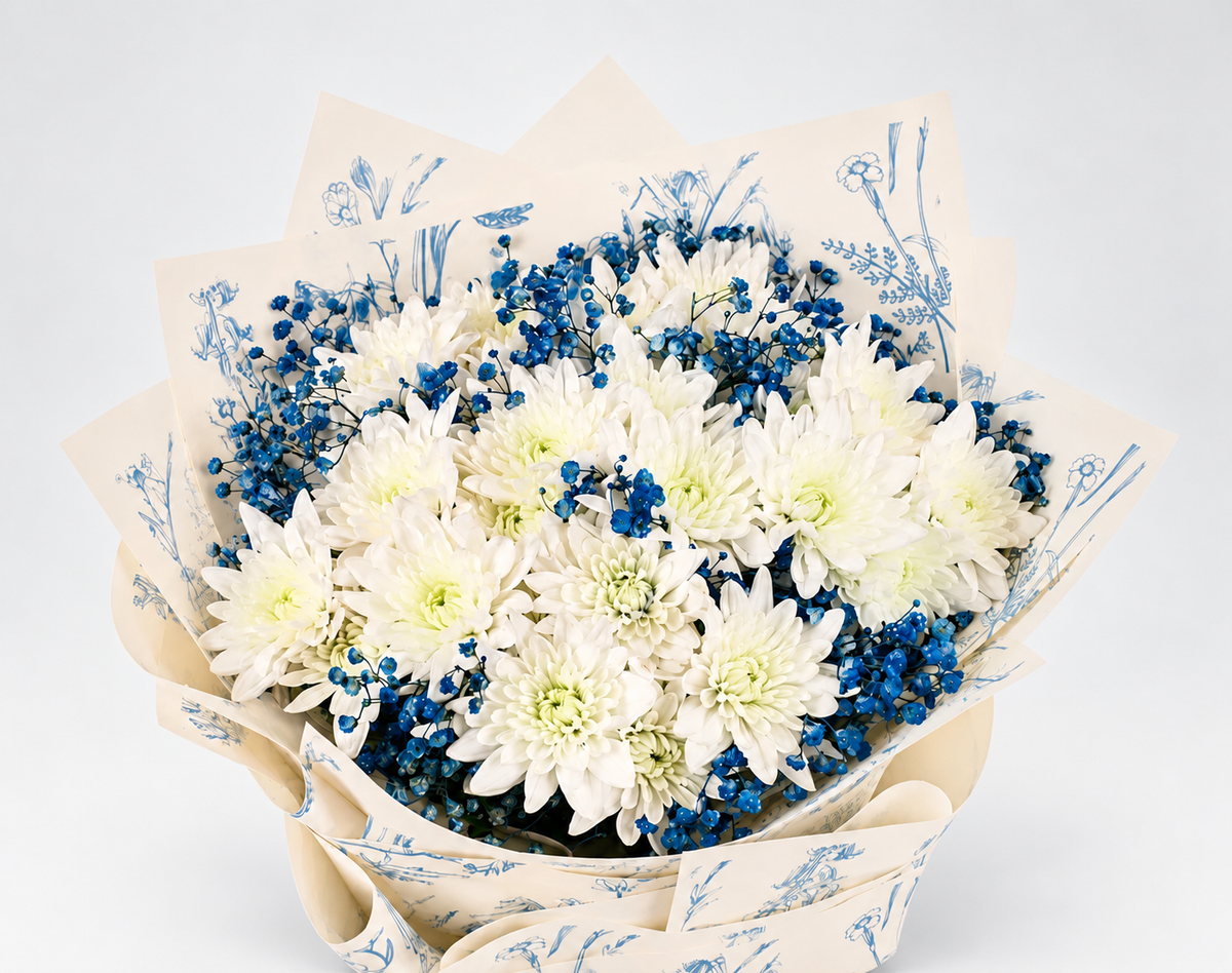 A bouquet of white and blue flowers wrapped in white paper with a blue botanical pattern and tied with a dark blue ribbon, displayed on a white pedestal against a white background.