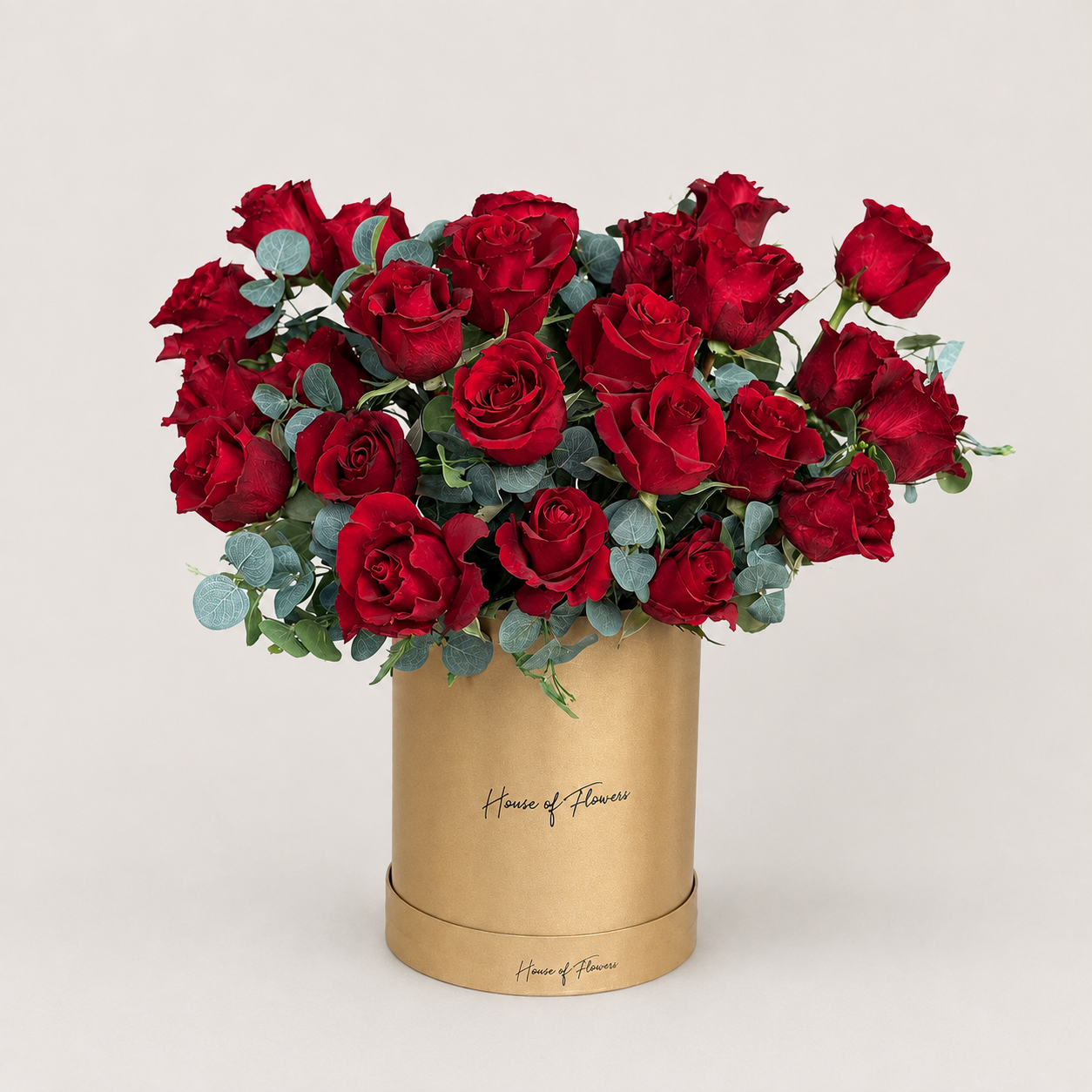 A bouquet of red roses with green eucalyptus stems arranged in a white cylindrical box printed with the gold script &