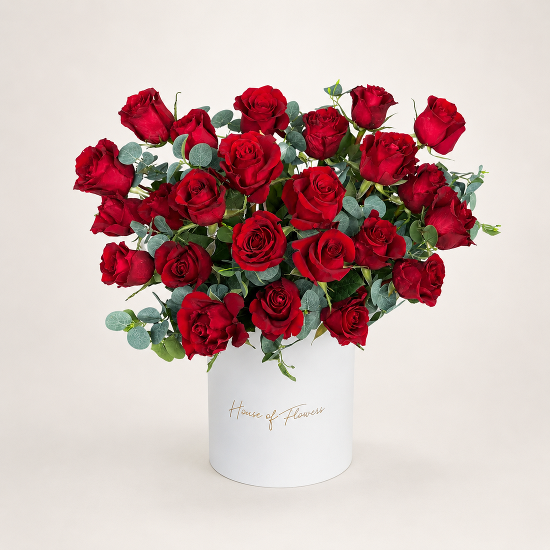 A bouquet of red roses with green eucalyptus stems arranged in a white cylindrical box printed with the gold script &