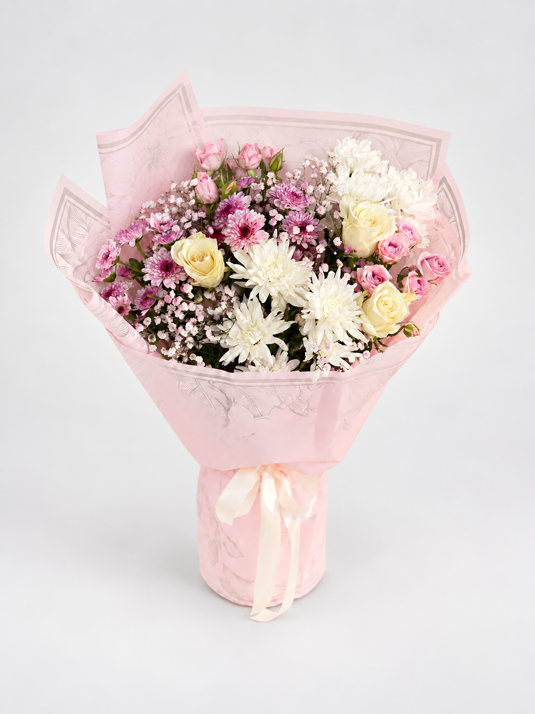 A floral bouquet wrapped in light pink tissue paper and tied with a pale pink ribbon, featuring white and pink flowers with small filler blossoms, photographed on a clean light gray studio background.