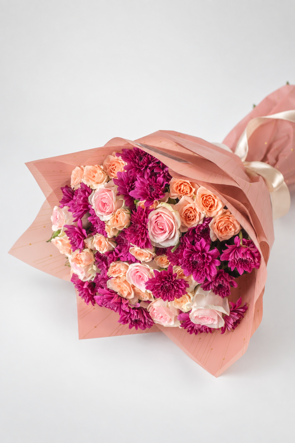 A bouquet of flowers consisting of roses and chrysanthemums in shades of pink and purple, wrapped in pink paper with a ribbon and a card that reads &