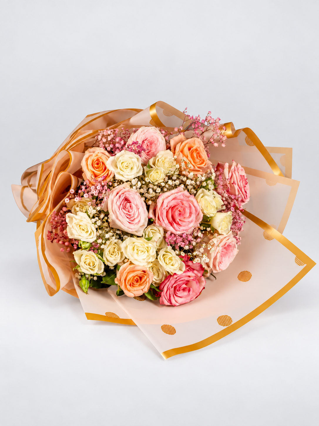 A bouquet of pink, peach, and white roses wrapped in light pink polka-dot paper with gold trim, tied with a satin ribbon, displayed on a cream pedestal against a white background.