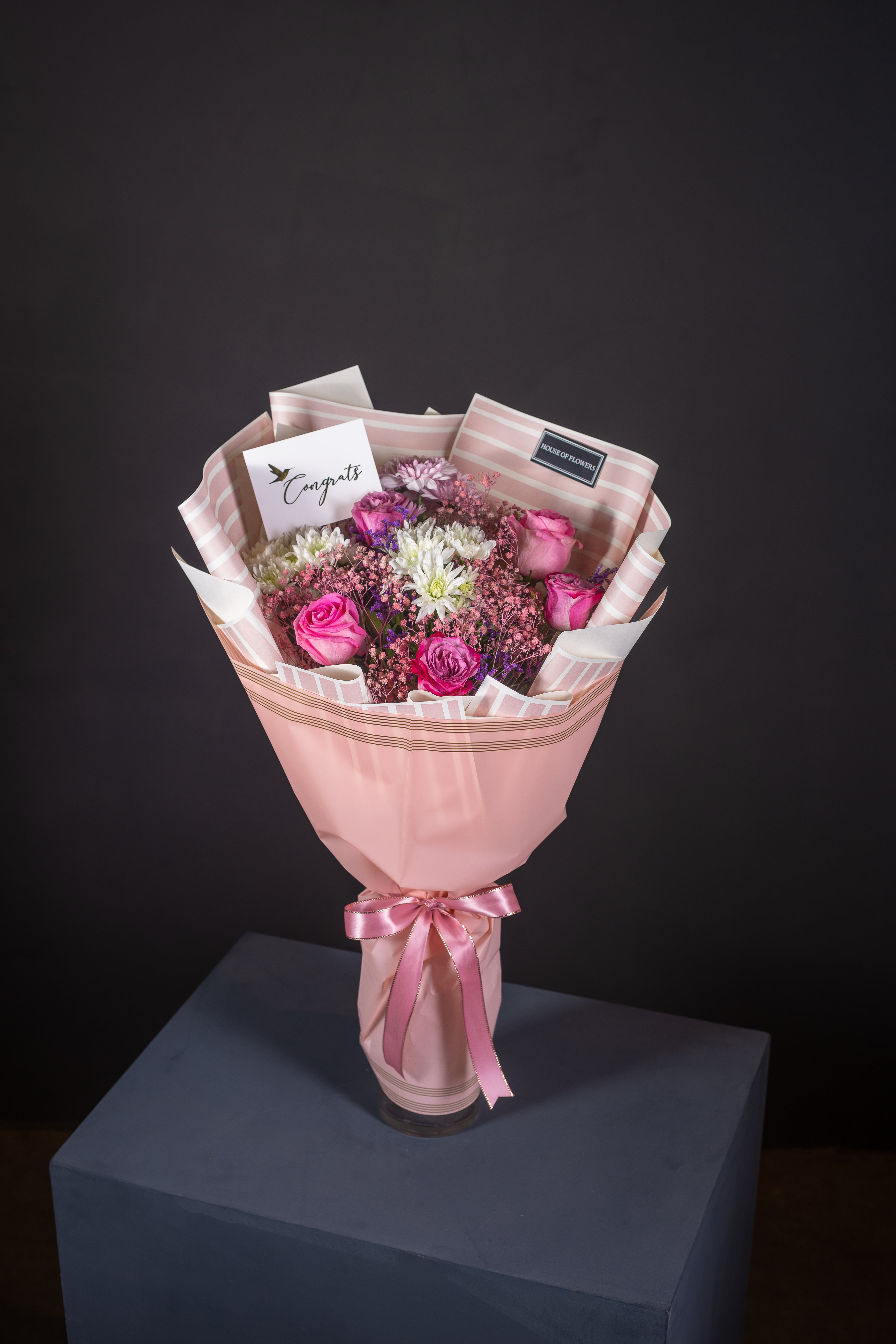 A bouquet of flowers including roses and chrysanthemums in purple, pink, and white colors, with a pink veil wrapped around the stems, tied with a pink ribbon.