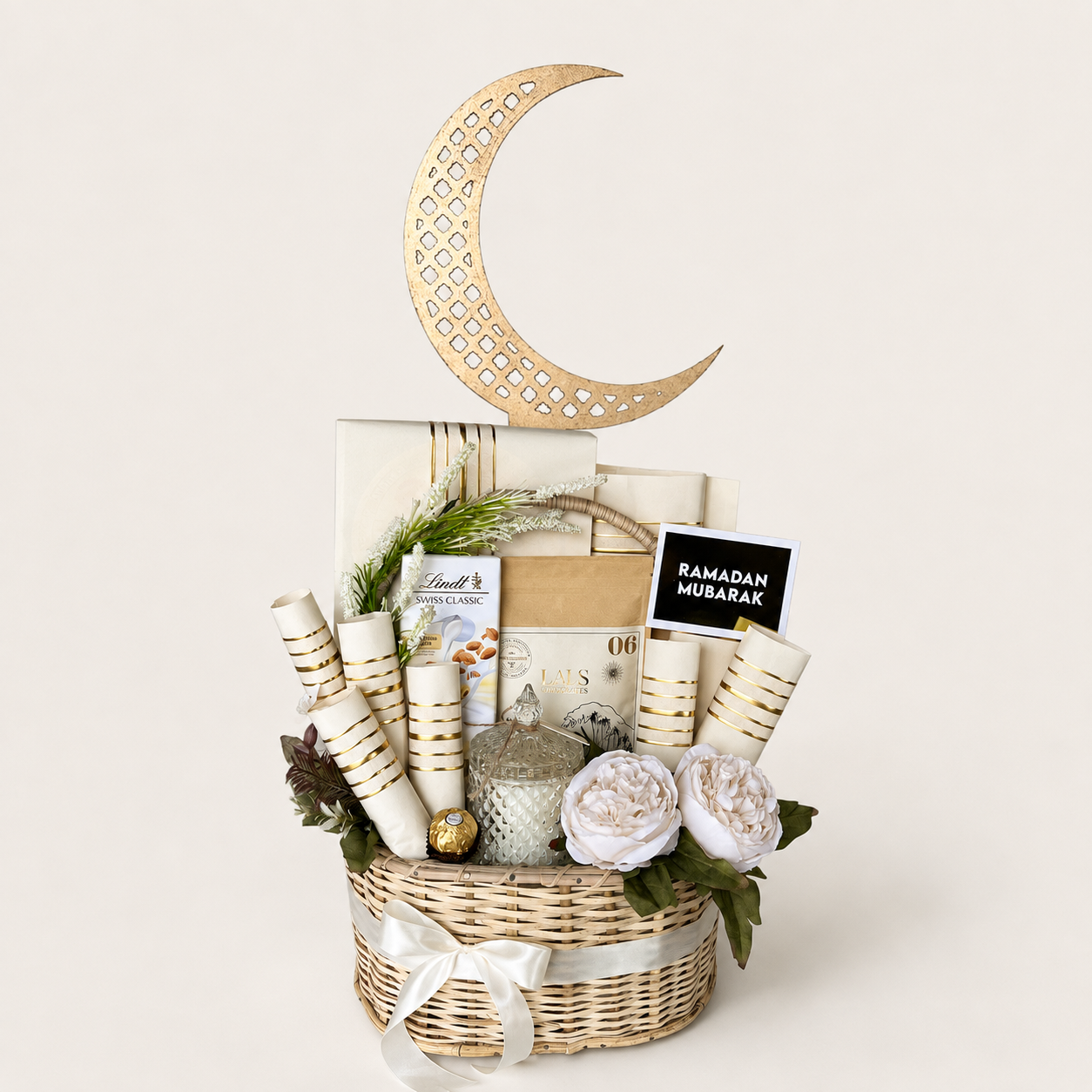 A Ramadan gift basket arrangement in a woven wicker basket, featuring a large gold crescent moon decoration, rolled white and gold mubarak cards, wrapped gift boxes, a small silver lantern, faux white flowers, and a black sign reading &