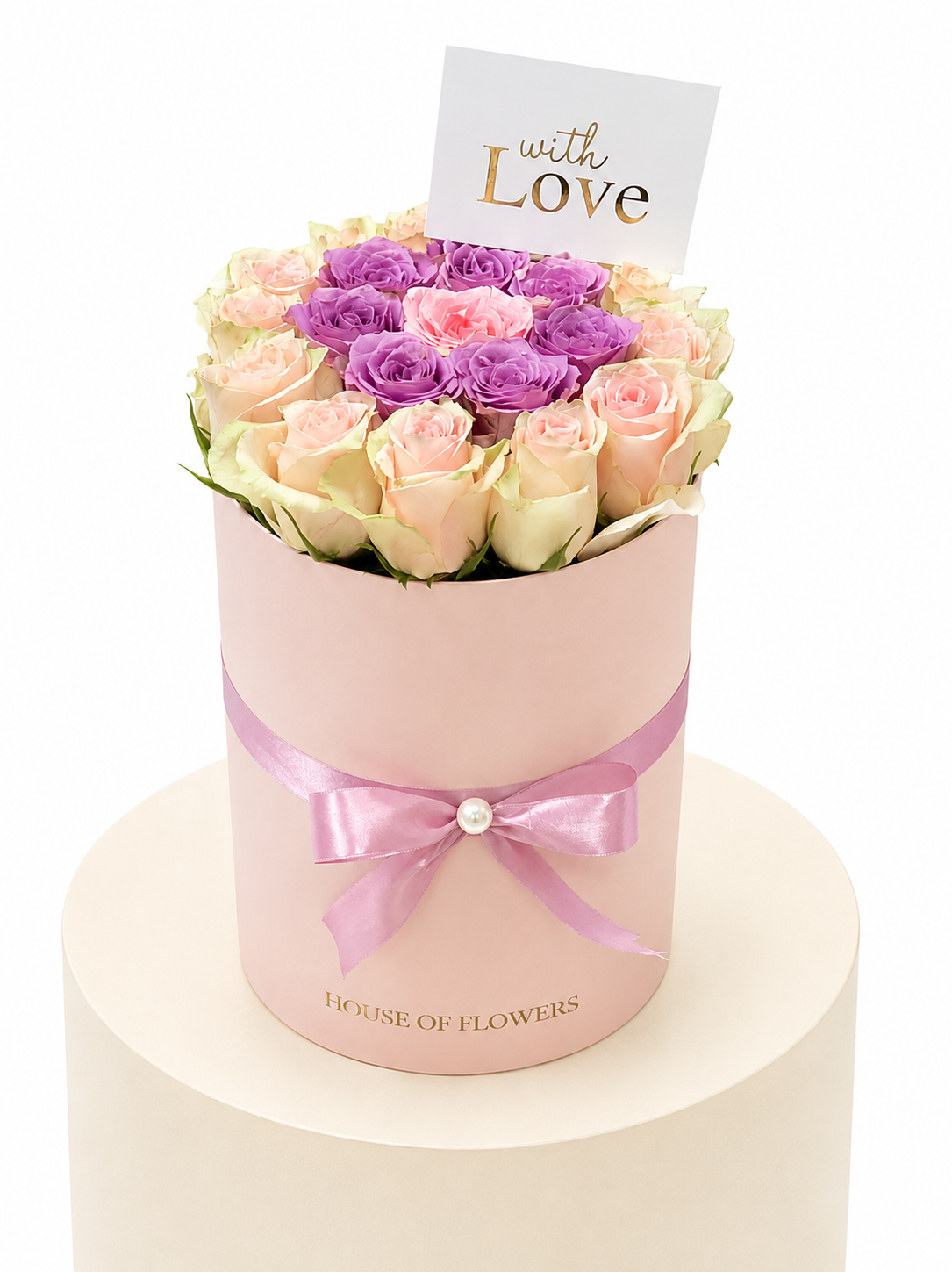 A round pink flower box filled with pale pink and purple roses, tied with a pink ribbon and topped with a small card that reads &