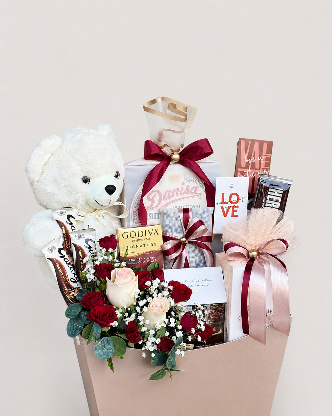 A curated gift basket arrangement in a blush pink bag, featuring a white teddy bear, a bouquet of red and blush roses with greenery, assorted chocolates including Godiva and Valhalla, and wrapped gift boxes tied with dark red and blush ribbons, set against a plain light beige background.