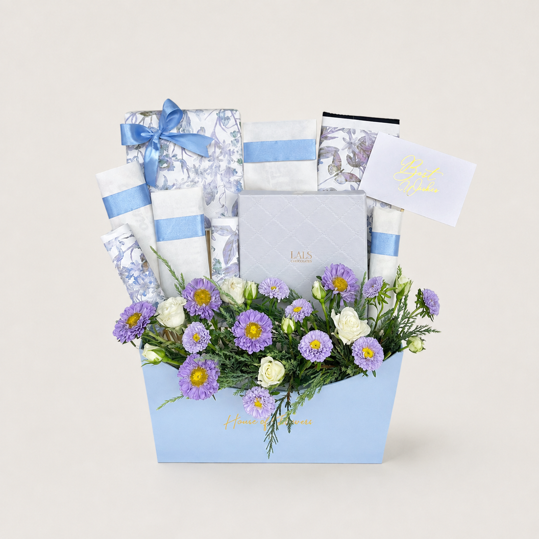 A baby gift basket arrangement in a light blue box, filled with baby wipes, a boxed item tied with a blue ribbon, a card, and a floral bouquet of purple and white flowers, on a light beige background.