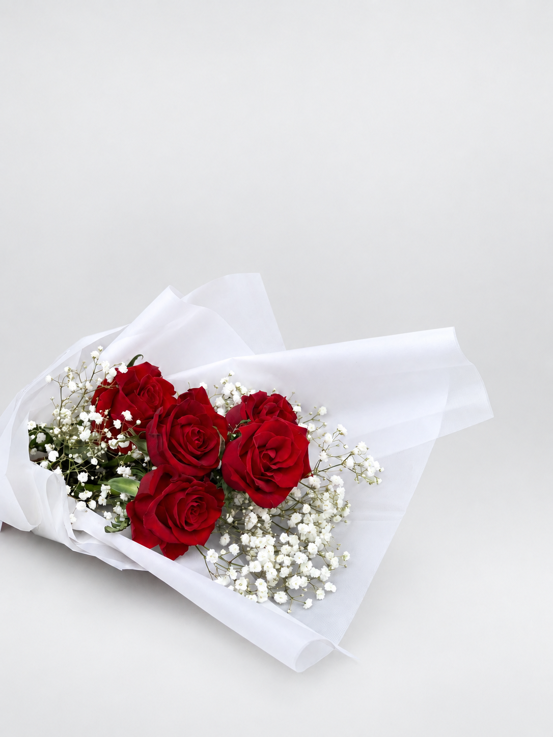 A bouquet of five red roses wrapped in white tissue paper with a dark red ribbon bow, displayed on a light beige pedestal against a white background.