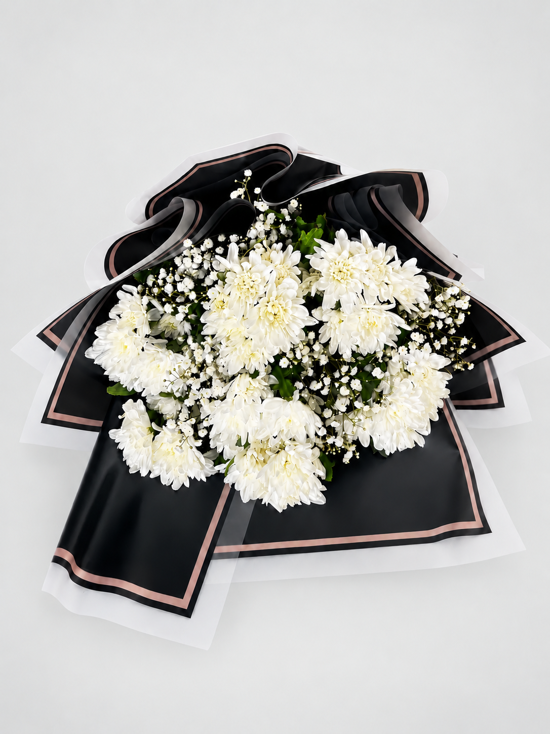 A bouquet of white chrysanthemums wrapped in black paper with white ribbon accents, displayed on a light beige pedestal against a white background.