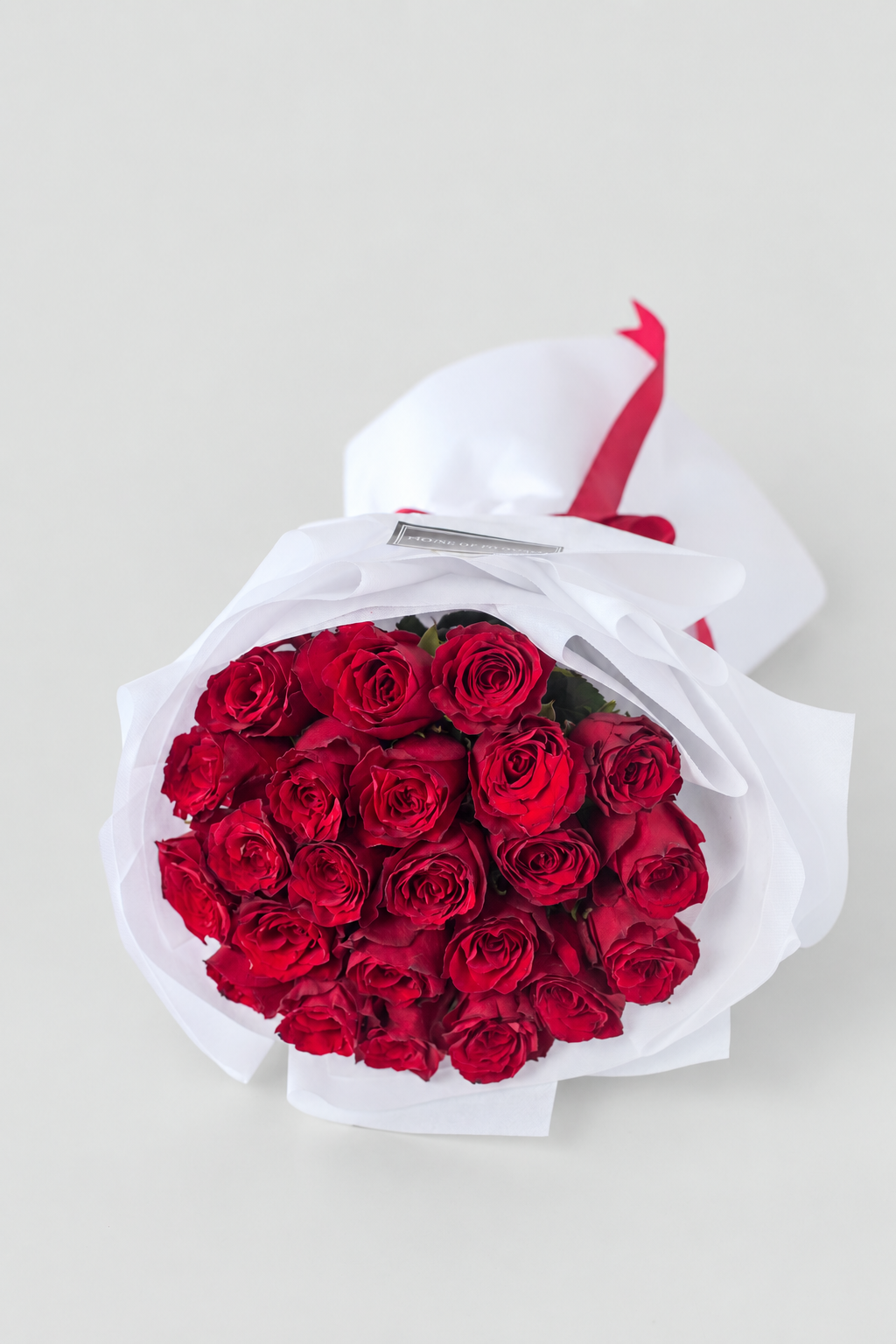 Bouquet of red roses wrapped in white paper with a red ribbon and a card on a white surface.
