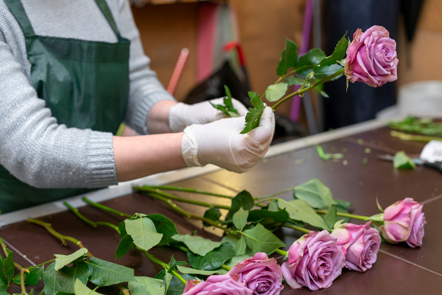 The photo shows a florist trimming the stems of purple roses for optimal care. To prolong their life, steps include choosing fresh roses, angled stem cuts for water uptake, leaf removal to hinder bacteria, and using a vase with water and flower food.