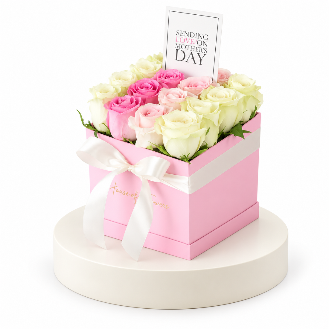 A pink square gift box filled with pink and cream roses, tied with a white ribbon and topped with a card that reads &