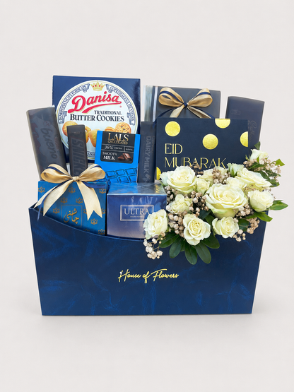 House of Flowers Eid Treats gift box with Danisa Butter Cookies, Lals Chocolates, Cadbury, J. ULTRA perfume, Mithai, and fresh white roses with gypsophila in a navy blue branded box