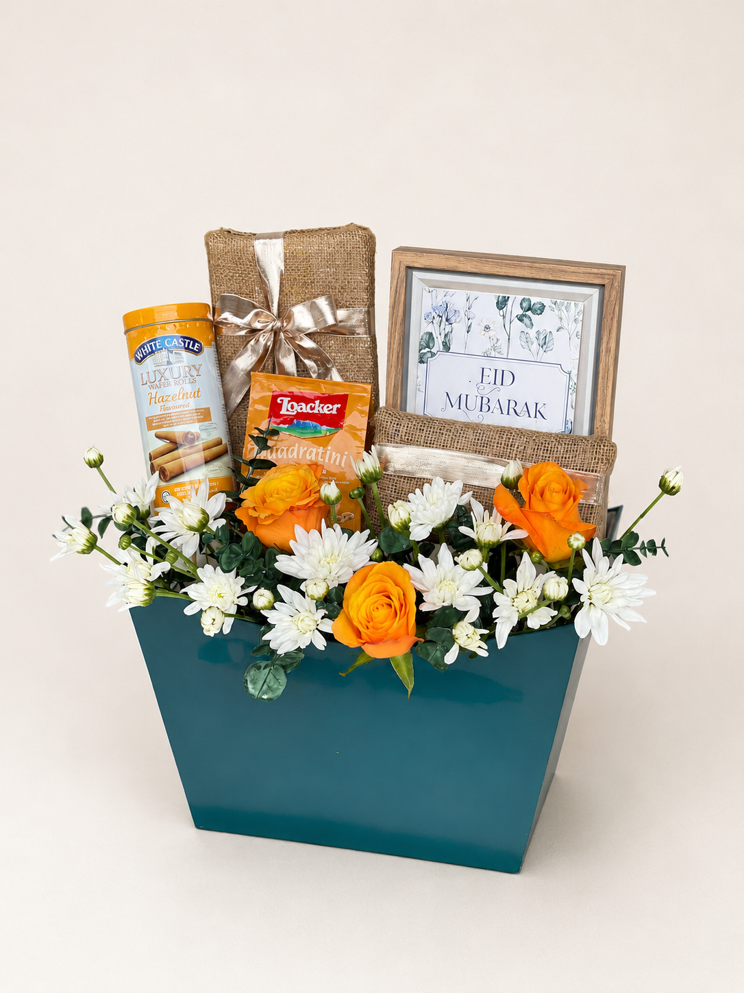 Close-up of Eid ul Adha Treats teal gift box by House of Flowers showing orange roses, white chrysanthemums, White Castle Luxury Hazelnut wafers, Loacker Quadratini wafers, jute-wrapped chocolates, and an Eid Mubarak framed print