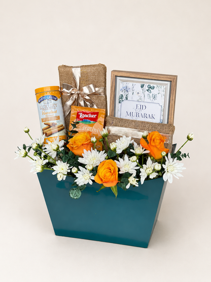 Close-up of Eid ul Adha Treats teal gift box by House of Flowers showing orange roses, white chrysanthemums, White Castle Luxury Hazelnut wafers, Loacker Quadratini wafers, jute-wrapped chocolates, and an Eid Mubarak framed print