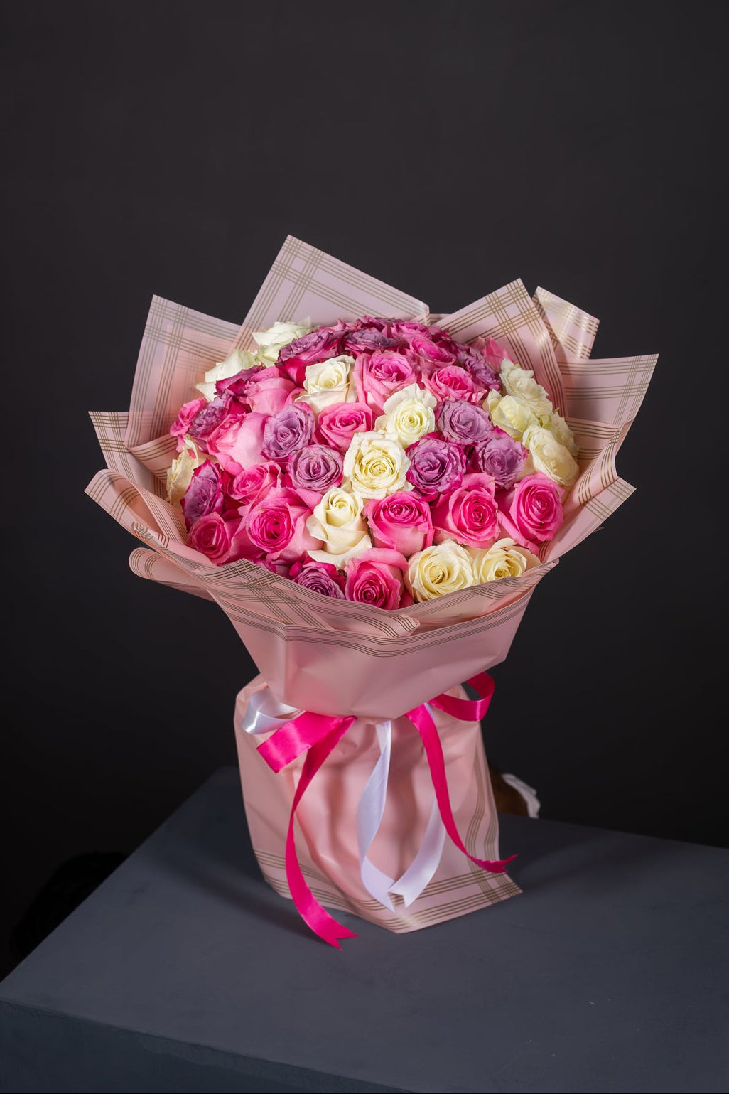 A bouquet of fresh roses, featuring a mix of pink, purple, and white roses, wrapped in yellow and clear paper with a pink ribbon.