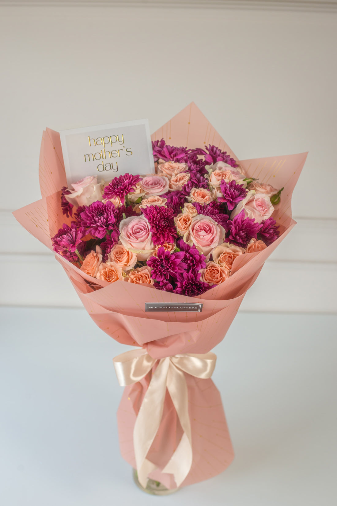 A bouquet of flowers consisting of roses and chrysanthemums in shades of pink and purple, wrapped in pink paper with a ribbon and a card that reads &