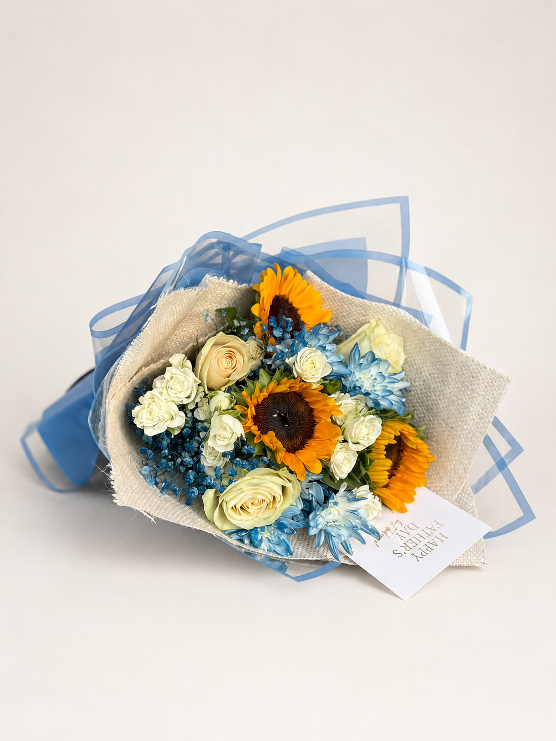 A dried flower bouquet featuring sunflowers, cream roses, blue chrysanthemums, and blue filler flowers, wrapped in a beige burlap-style paper sleeve with a sheer blue ribbon and a small white card, photographed on a light beige background.