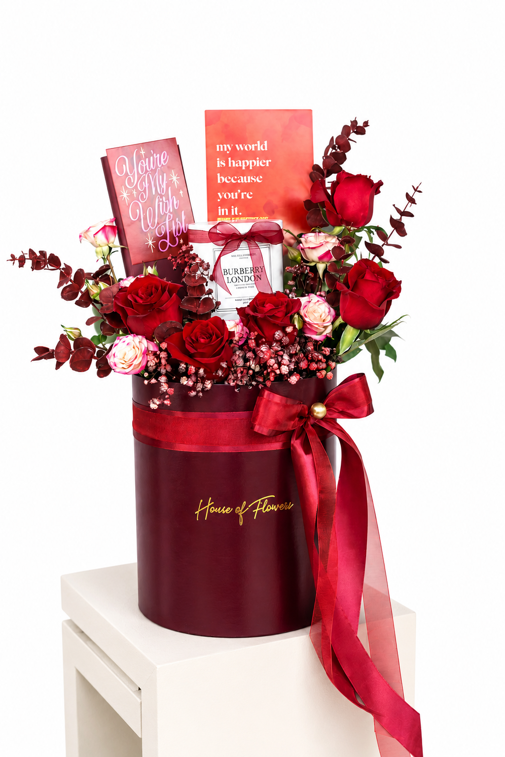 A red and pink floral gift box arrangement from House of Flowers, featuring red roses, pink blossoms, greenery, two greeting cards, and a small bottle, displayed on a white pedestal against a white background.