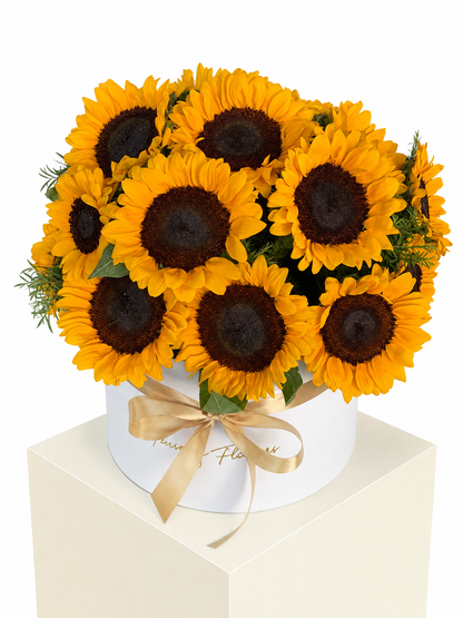A bouquet of bright yellow sunflowers with dark brown centers arranged in a round white box tied with a gold ribbon, displayed on a white pedestal against a white background.