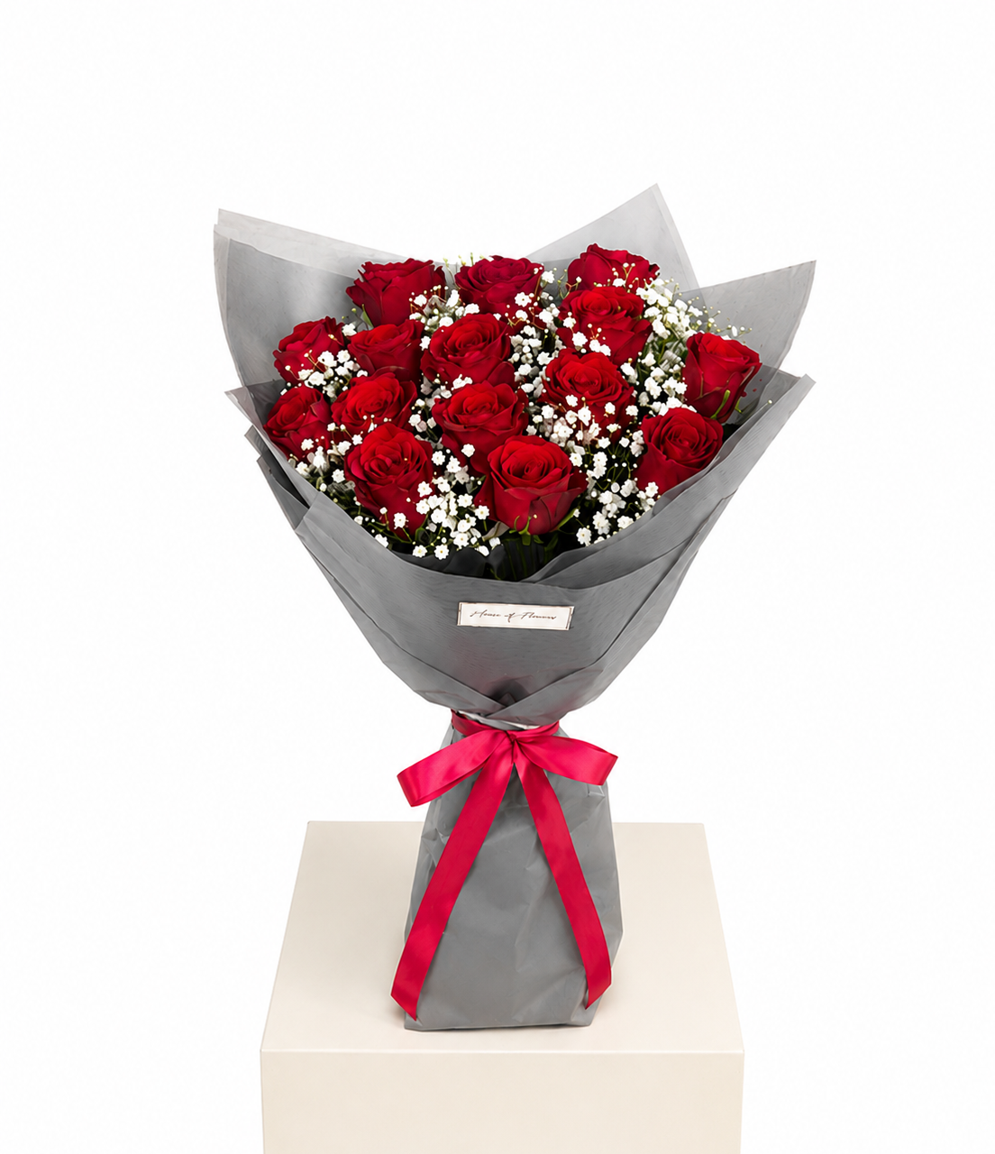 A bouquet of 12 red roses wrapped in gray paper with a bright red ribbon bow, displayed on a white pedestal against a white background.