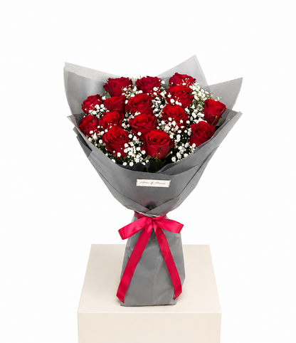 A bouquet of 12 red roses wrapped in gray paper with a bright red ribbon bow, displayed on a white pedestal against a white background.