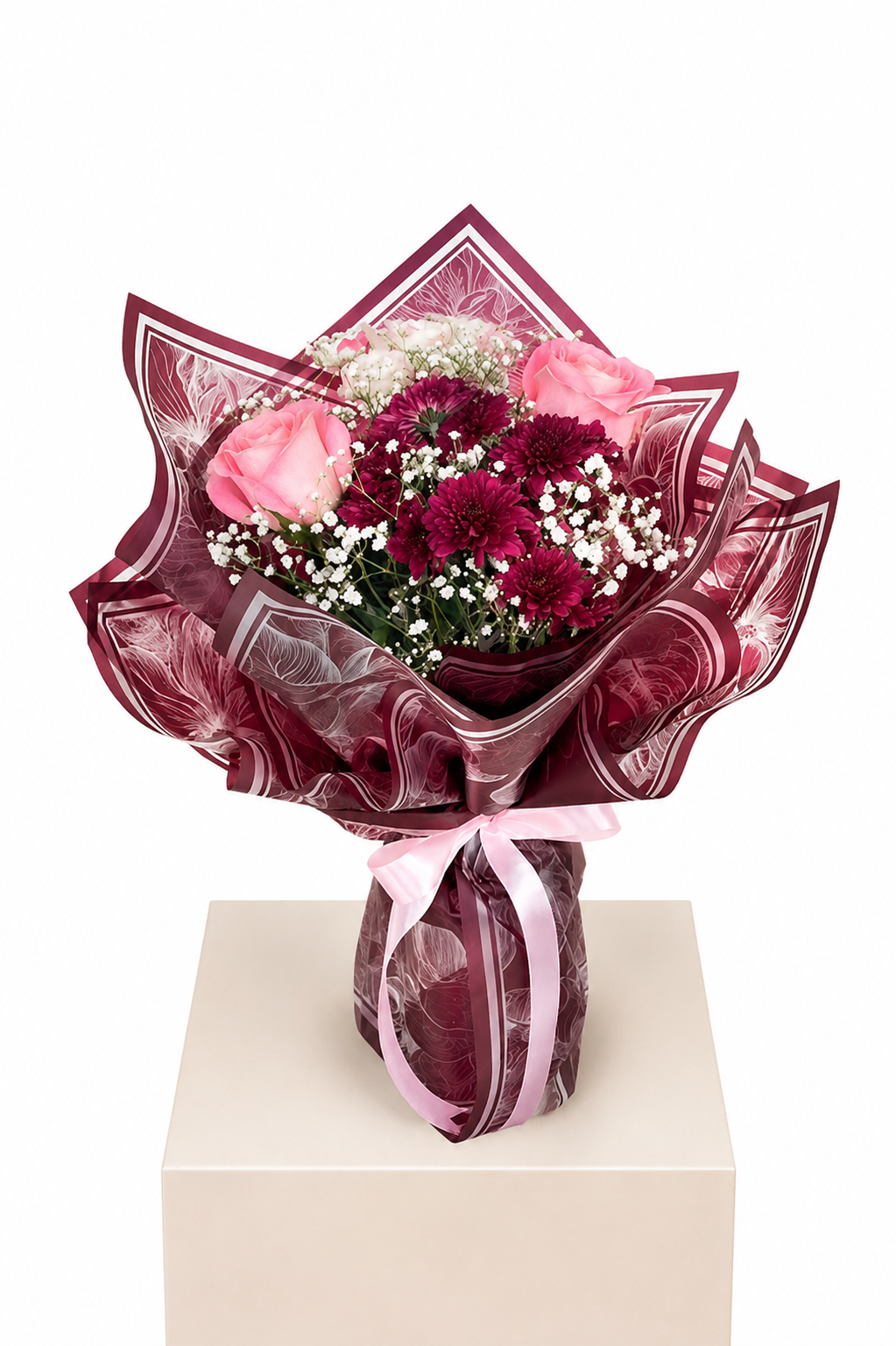 A floral bouquet wrapped in shiny dark burgundy paper and tied with a pink ribbon, displayed on a beige pedestal against a white background. The bouquet includes pink roses, deep burgundy chrysanthemums, and white baby&