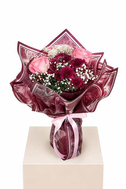 A floral bouquet wrapped in shiny dark burgundy paper and tied with a pink ribbon, displayed on a beige pedestal against a white background. The bouquet includes pink roses, deep burgundy chrysanthemums, and white baby&