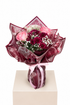 A floral bouquet wrapped in shiny dark burgundy paper and tied with a pink ribbon, displayed on a beige pedestal against a white background. The bouquet includes pink roses, deep burgundy chrysanthemums, and white baby&