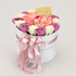 A round white gift box filled with pink, peach, cream, and purple roses, tied with a large pale pink satin bow. A small gold card reading &