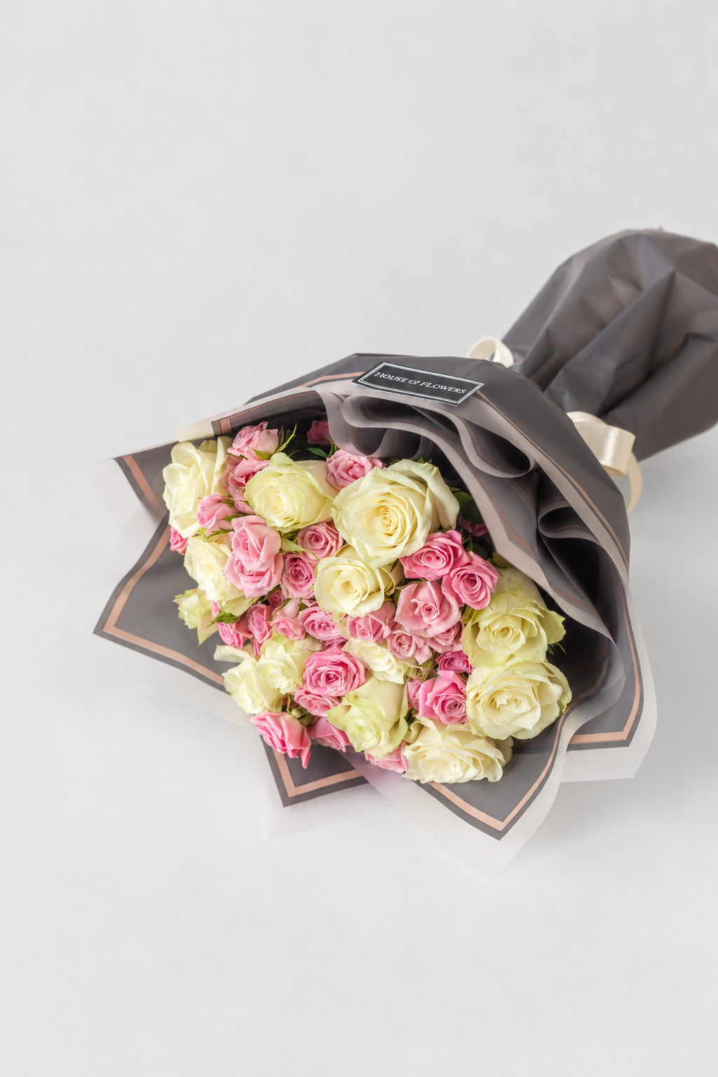 A bouquet of mixed pink and pale yellow roses wrapped in dark gray paper with a small black label, photographed on a clean light gray background.