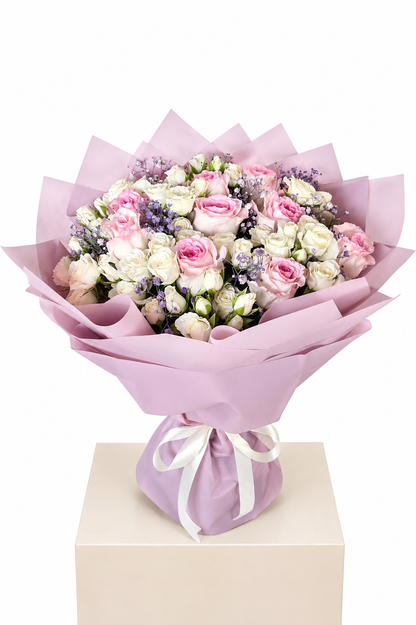 A bouquet of pink and white roses with small purple filler flowers, wrapped in light pink tissue paper and tied with a white ribbon, displayed on a beige pedestal against a white background.
