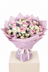 A bouquet of pink and white roses with small purple filler flowers, wrapped in light pink tissue paper and tied with a white ribbon, displayed on a beige pedestal against a white background.