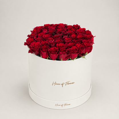 A round white gift box filled with a dense arrangement of deep red roses, with the brand name &
