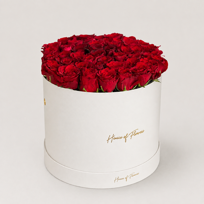 A bouquet of red roses with green eucalyptus stems arranged in a white cylindrical box printed with the gold script &