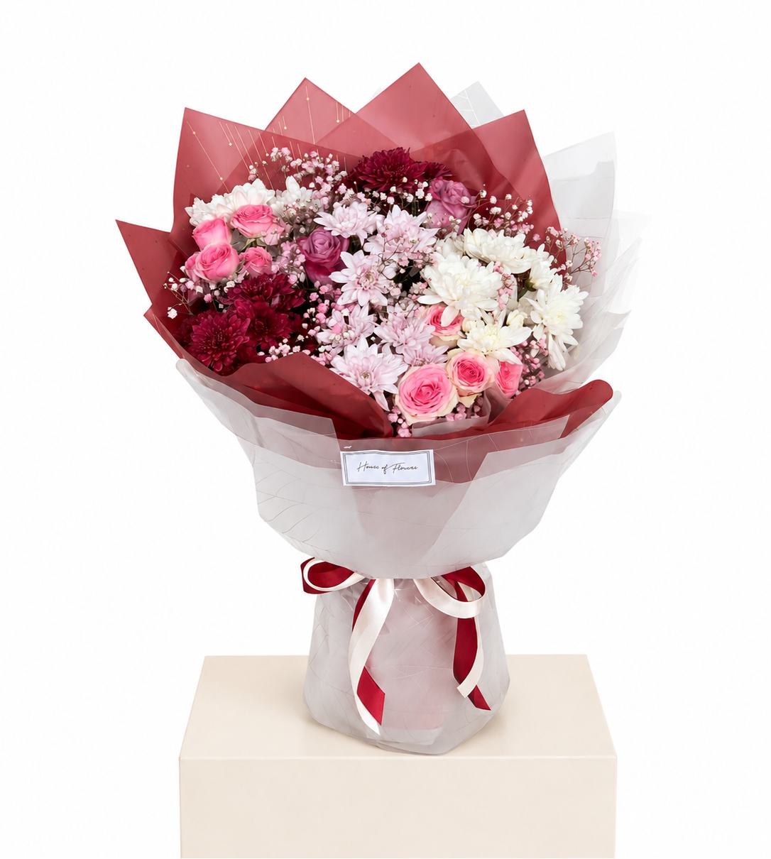 A bouquet of pink, red, and white flowers wrapped in red and white tissue paper, tied with a red ribbon, displayed on a beige pedestal against a white background.