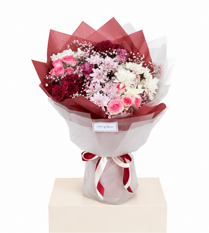A bouquet of pink, red, and white flowers wrapped in red and white tissue paper, tied with a red ribbon, displayed on a beige pedestal against a white background.