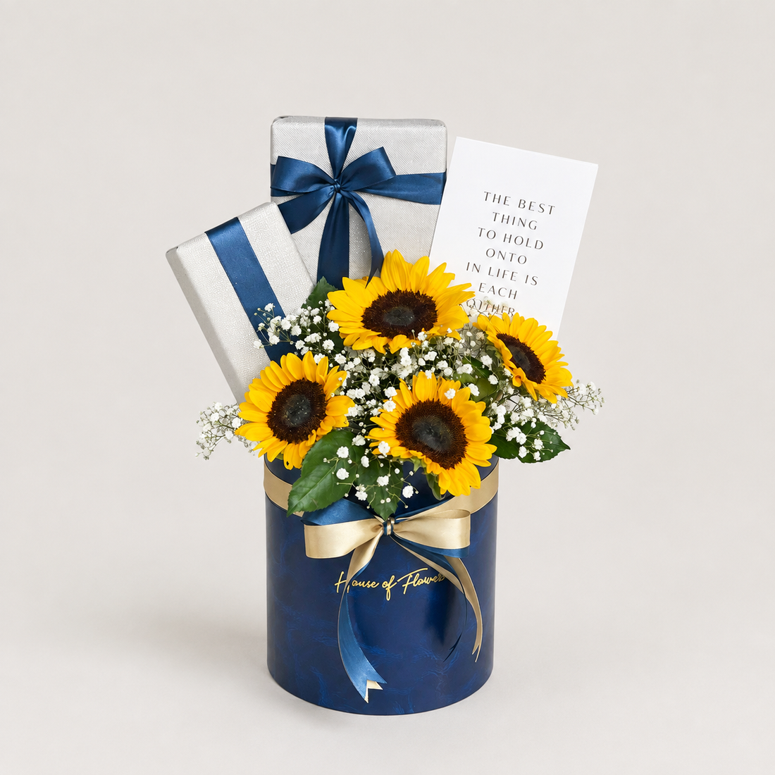 Sunflower gift arrangement in a dark blue cylindrical box with a gold ribbon, two wrapped gift boxes, and a printed card on a light neutral background.