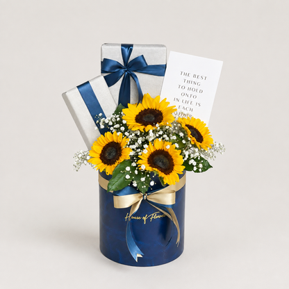 Sunflower gift arrangement in a dark blue cylindrical box with a gold ribbon, two wrapped gift boxes, and a printed card on a light neutral background.