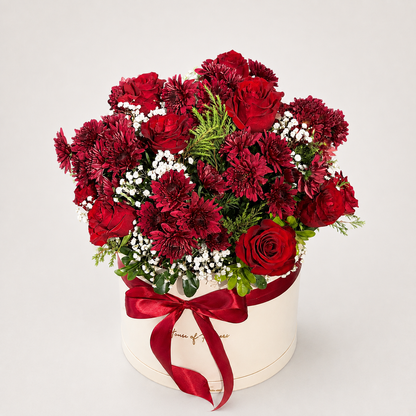 A round cream-colored flower box tied with a red ribbon, filled with a bouquet of red roses, dark red chrysanthemums, burgundy carnations, and white baby&