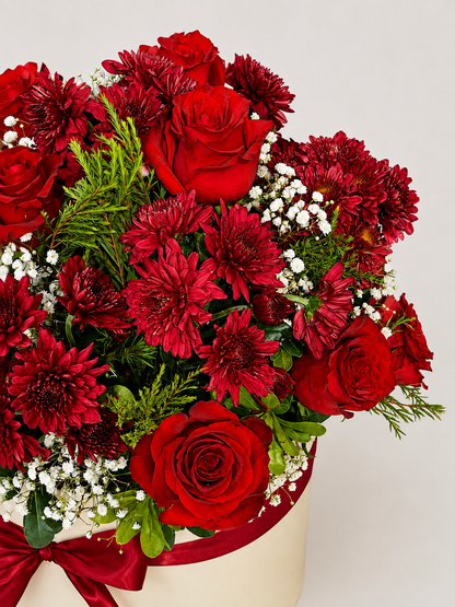 A round cream-colored flower box tied with a red ribbon, filled with a bouquet of red roses, dark red chrysanthemums, burgundy carnations, and white baby&