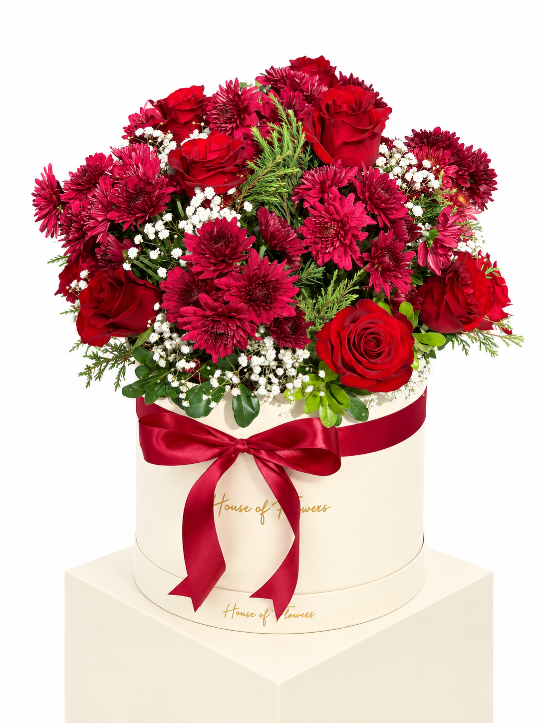 A round cream-colored flower box tied with a red ribbon, filled with a bouquet of red roses, dark red chrysanthemums, burgundy carnations, and white baby&