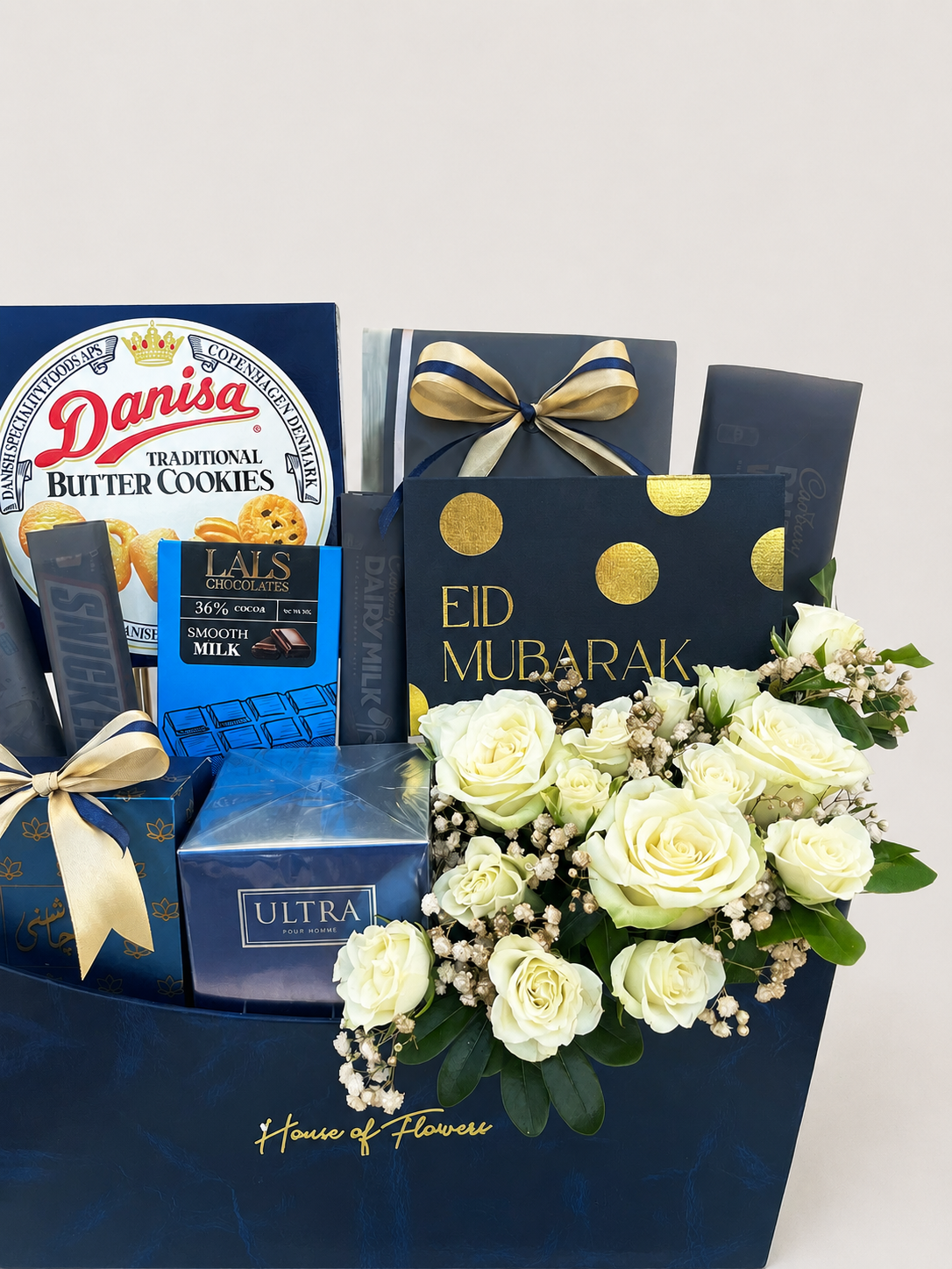 House of Flowers Eid Treats gift box with Danisa Butter Cookies, Lals Chocolates, Cadbury, J. ULTRA perfume, Mithai, and fresh white roses with gypsophila in a navy blue branded box