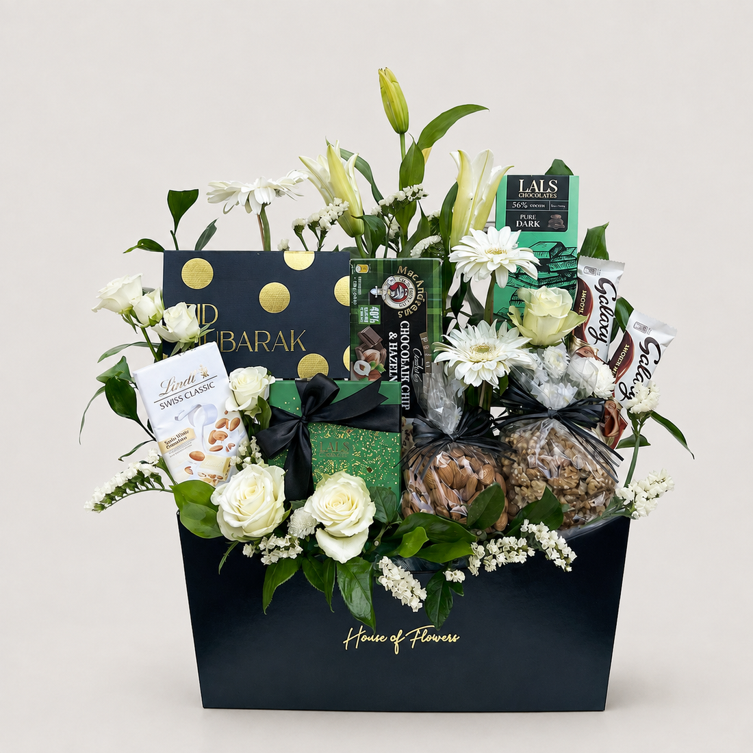 A luxury gourmet gift basket from House of Flowers, featuring a black box filled with white roses, green lilies, white daisies, and assorted gourmet treats including chocolate, crackers, and sweets. The basket is decorated with black and gold ribbons and branded packaging.